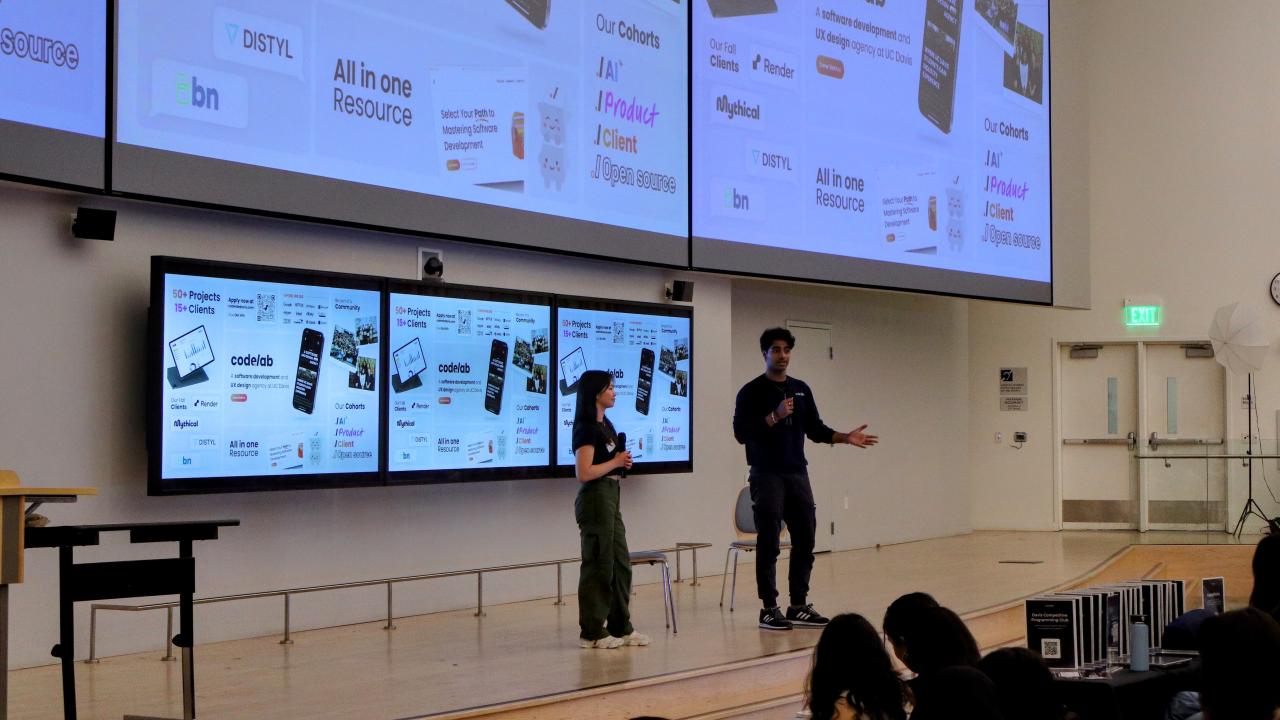 Two students speaking into microphones in front of giant screens