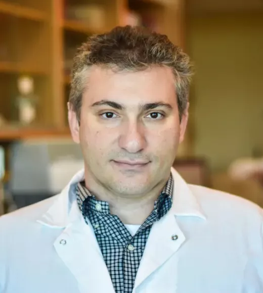 A man with salt-and-pepper hair wearing a lab coat looks straight into the camera