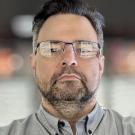 Kristian Stevens with glasses and a beard wearing a gray button-up shirt.